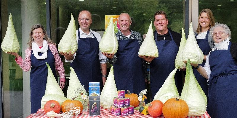 Sechs Personen in blauen Schürzen halten große Spitzkohlköpfe hoch, vor ihnen ein Tisch mit Kürbissen, Krautdosen und Deko.