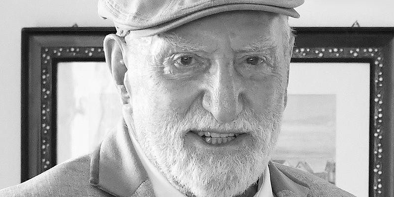 Schwarz-weiß-Foto eines älteren Mannes mit Bart, Flatcap und Anzug, der in die Kamera blickt.