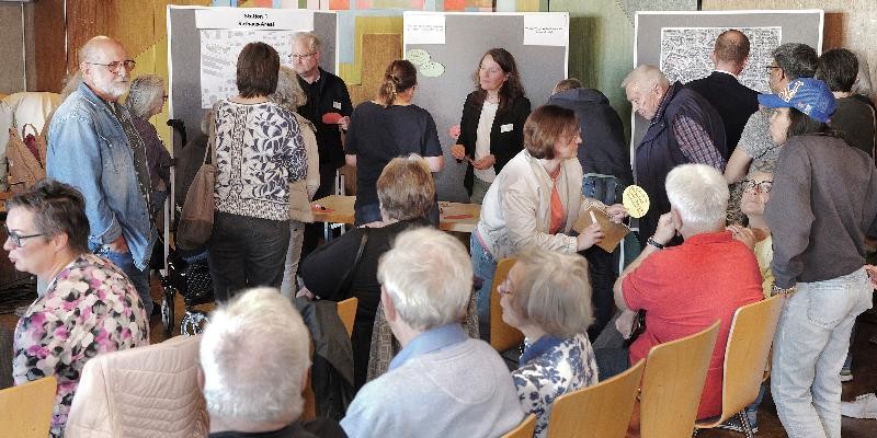 Menschen diskutieren bei einer Bürgerbeteiligung in kleinen Gruppen vor Stellwänden und Karten. Die Veranstaltung wirkt lebendig und gemeinschaftlich, viele Teilnehmer tauschen Ideen und Meinungen aus.