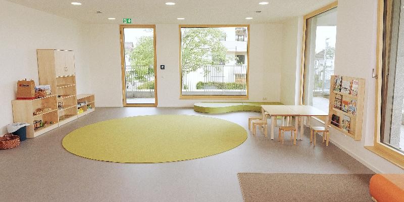 Heller Gruppenraum mit Spielteppich, Tisch und Regalen