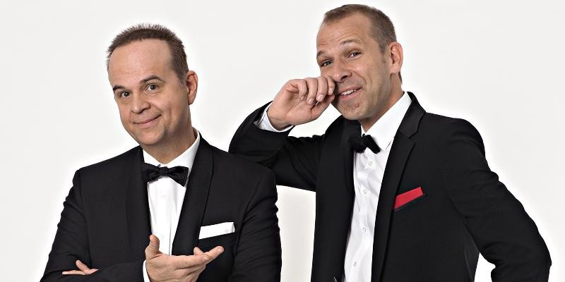 Zwei Männer im Smoking posieren humorvoll vor hellem Hintergrund und schauen lächelnd direkt in die Kamera.