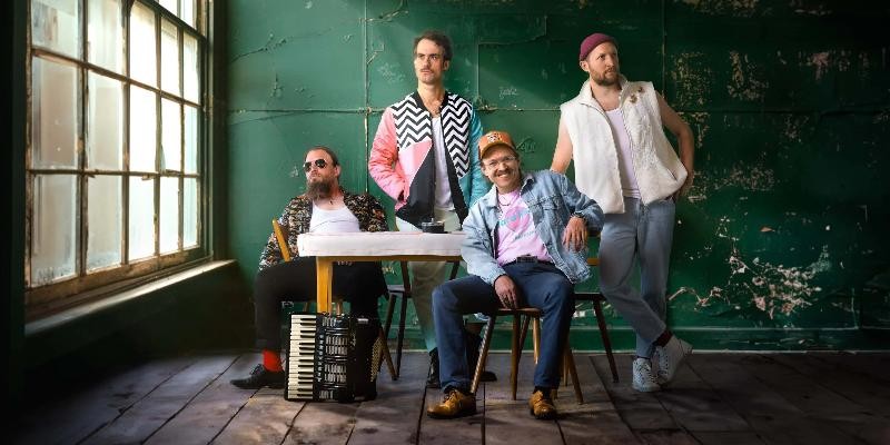 Vier Musiker posieren lässig in industriellem Raum mit Akkordeon, bunten Outfits und Vintage-Atmosphäre vor grüner Wand.