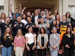 Gruppenbild neuer Azubis vor Rathaus: junge Erwachsene in Alltags- und Arbeitskleidung, lächeln, heben motiviert die Fäuste.