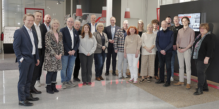 Eine Gruppe von rund zwanzig Personen posiert in einem modernen Innenraum für ein gemeinsames Foto, im Hintergrund sind Roto‑Schilder zu sehen.
