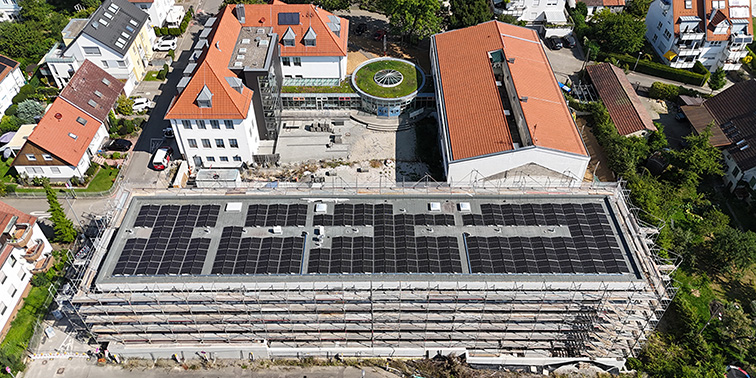 Luftaufnahme eines großen Gebäudes mit Solaranlage auf Dach, Baugerüst und umliegende Wohnhäuser sichtbar.