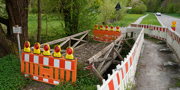 Eine gesperrte Holzbrücke wird von rot‑weißen Absperrungen und gelben Warnleuchten flankiert, daneben verläuft eine Straße durch grünes Gelände.