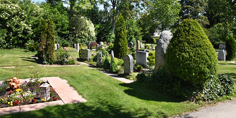Ein gepflegter Friedhof mit Grabsteinen, Blumenbeeten und Bäumen in ruhiger, grüner Umgebung.