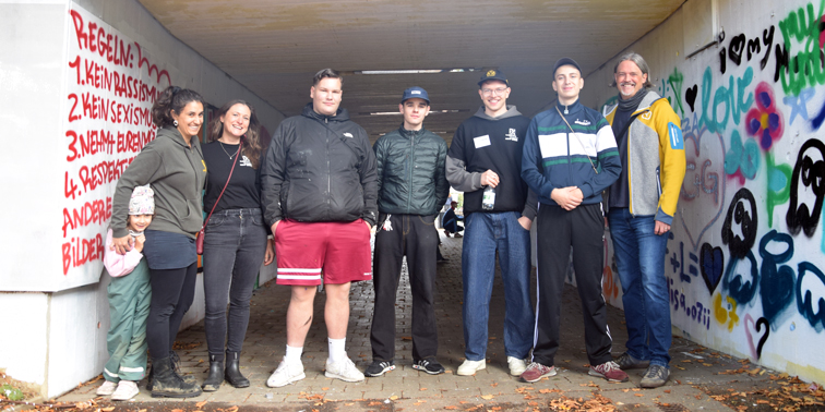 Gruppenfoto von acht Personen in einer Unterführung mit Regeln an der linken und bunten Graffiti an der rechten Wand.