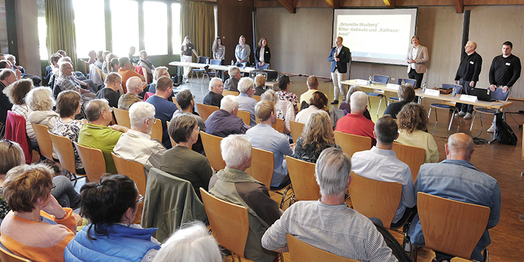 Eine größere Gruppe sitzt in einem Saal und verfolgt eine Präsentation, während mehrere Personen vorne vor einer Leinwand stehen.