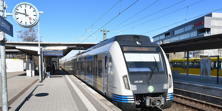 Das Bild zeigt eine S-Bahn am Bahnsteig eines modernen Bahnhofs bei sonnigem Wetter. Die Uhr zeigt etwa 11:42 Uhr. Im Hintergrund sind das Bosch-Gebäude und weitere Bahnsteige zu sehen.