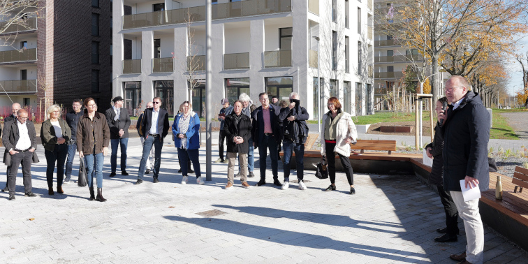 Gruppe von Menschen steht bei Sonnenschein auf einem Platz vor modernen Wohngebäuden, der Oberbürgermeister spricht mit Mikrofon