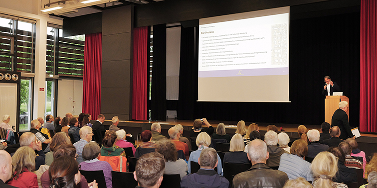 Ein voll besetzter Saal, in dem viele Menschen einer Präsentation auf einer Bühne mit Leinwand folgen.