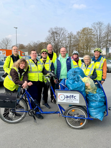 Gruppe in Warnwesten steht mit Fahrrädern und Anhänger voller Müllsäcke bei einer Putzaktion.