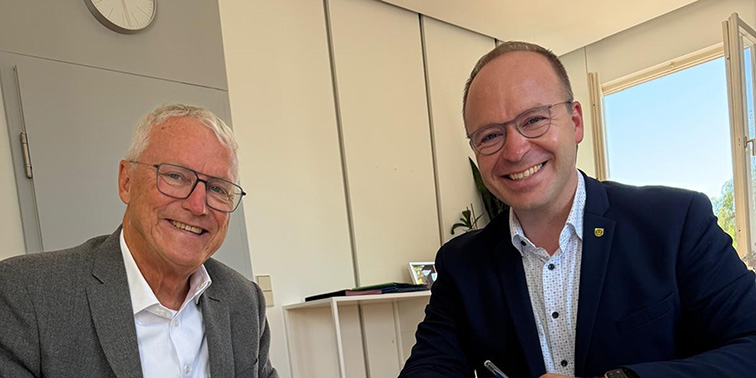 Zwei Männer in Businesskleidung sitzen lächelnd an einem Tisch und unterzeichnen ein Dokument in einem hellen Büro.