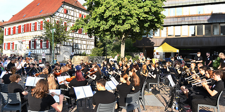 Ein großes Orchester spielt im Freien auf einem Platz, umgeben von Publikum. Die Musiker tragen schwarze Kleidung und spielen verschiedene Instrumente, darunter Geigen, Trompeten und Flöten. Im Hintergrund sind Fachwerkhäuser und weitere Gebäude zu sehen.