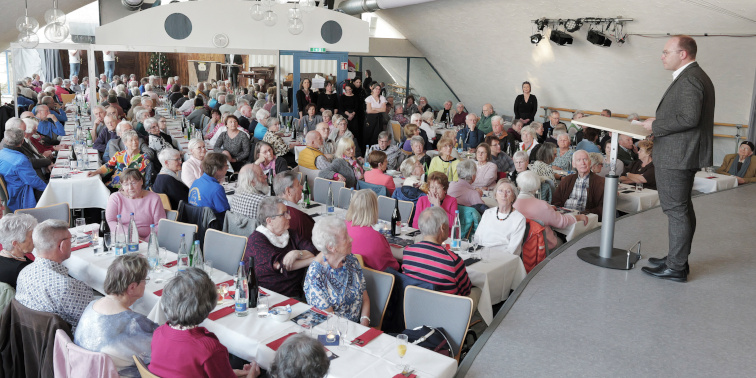 OB Ruppaner spricht auf einer Bühne vor vielen Seniorinnen und Senioren, die an Tischen sitzen.