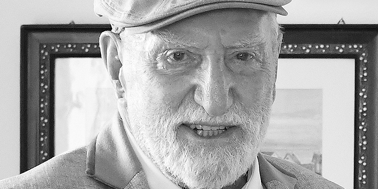 Schwarz-weiß-Foto eines älteren Mannes mit Bart, Flatcap und Anzug, der in die Kamera blickt.