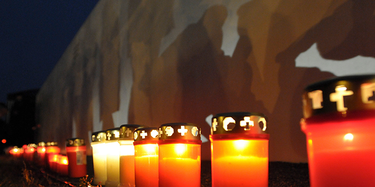 Gedenkstiftung schreibt Projektpreis aus Reihe leuchtender Grabkerzen bei Nacht vor einer Wand mit Schatten von Menschen