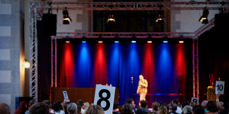 Publikum hält Bewertungskarten hoch, auf einer beleuchteten Bühne tritt ein Comedian vor rotem und blauem Licht auf.