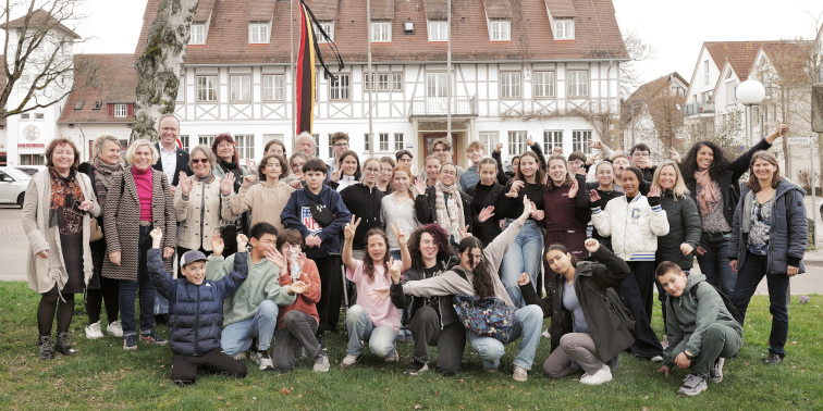 Große Gruppe von Jugendlichen und Erwachsenen posiert vor einem Gebäude mit Fahnen und lächelt in die Kamera.