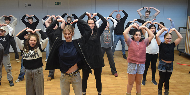 Eine Gruppe von Kindern und Erwachsenen formt gemeinsam Herzgesten in einem Bewegungsraum.