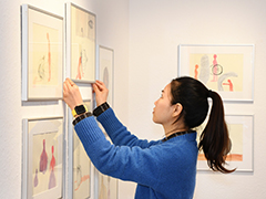 Eine Frau in blauem Pullover hängt in einer Galerie Bilder an die Wand. Die Bilder zeigen abstrakte Zeichnungen in hellen Farben.