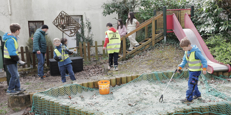 Kinder in Warnwesten sammeln Müll auf einem Spielplatz neben Rutsche und Sandfläche.