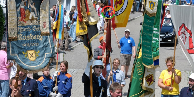Vereinsmitglieder in Tracht, mit Vereinsfahnen und Besucher bei Festumzug in einer Musberger Straße.