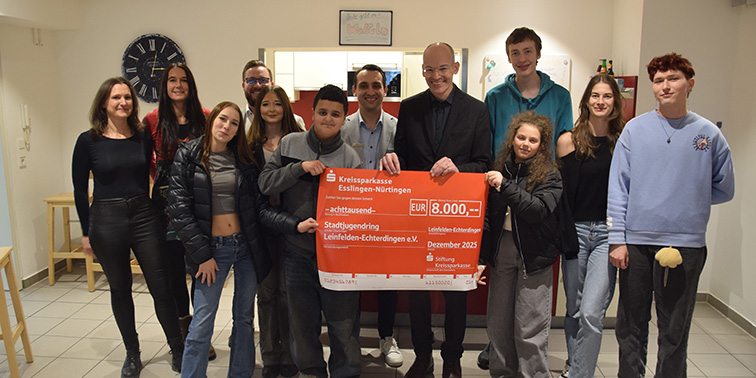 Gruppe von Jugendlichen und Erwachsenen steht in einem Raum und hält gemeinsam einen Spendenscheck über 8.000 Euro der Kreissparkasse.