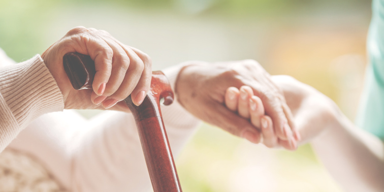 Eine ältere Person hält einen Gehstock, während eine helfende Hand sie fürsorglich unterstützt.