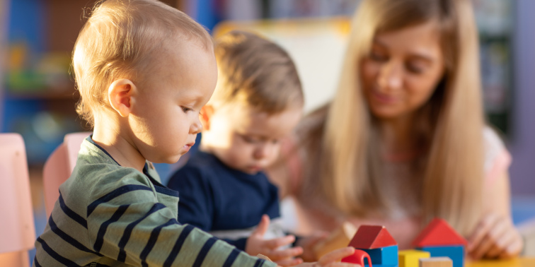 Zwei Kleinkinder spielen konzentriert mit bunten Holzklötzen, im Hintergrund schaut eine Erzieherin lächelnd zu.