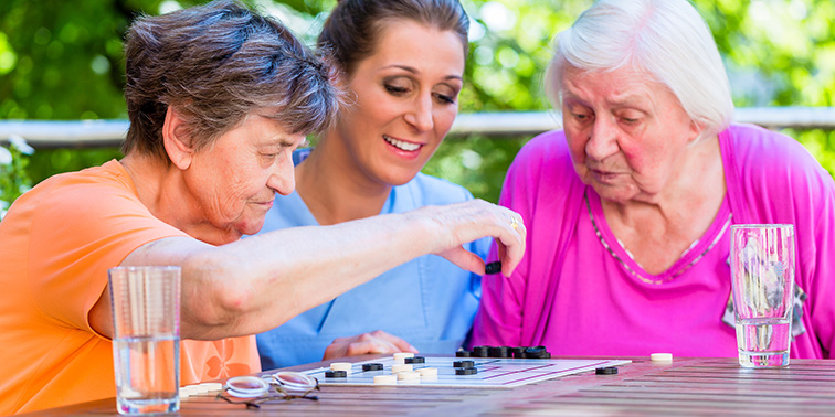 Eine Pflegekraft spielt mit zwei älteren Personen im Freien ein Brettspiel an einem Holztisch.