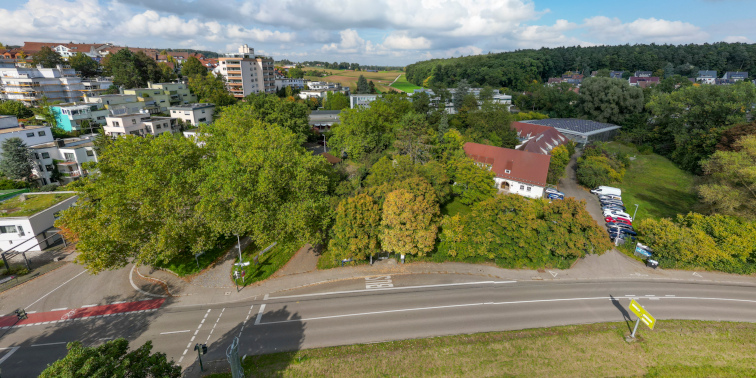 Luftaufnahme einer Straße mit Abzweigung, umgeben von Bäumen, Wohnhäusern und einem größeren Gebäude mit rotem Dach und Parkplatz.