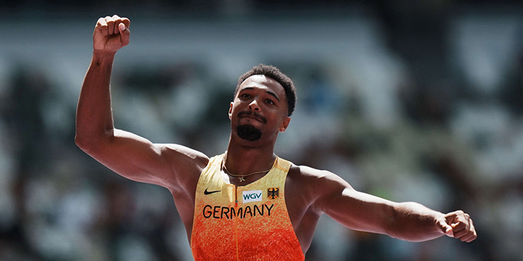 Ein Leichtathlet im orange-gelben Trikot mit der Aufschrift „Germany“ jubelt auf der Laufbahn mit erhobenem Arm im Stadion.