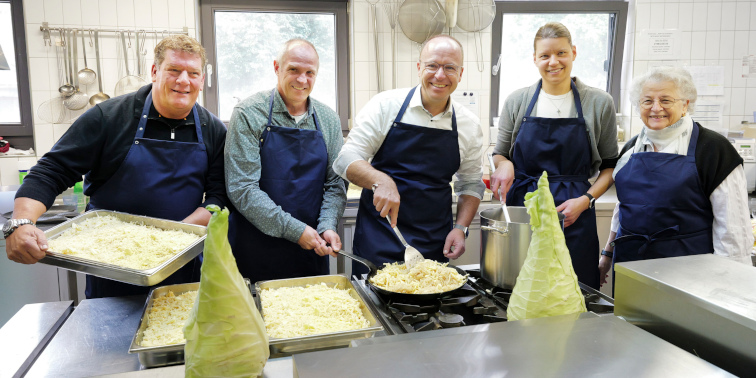 Fünf Personen in blauen Schürzen kochen gemeinsam in einer Großküche, lachen und bereiten Kraut und Nudeln in großen Pfannen zu.
