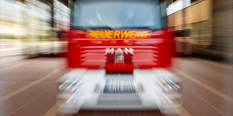 Rotes Feuerwehrfahrzeug mit der Aufschrift „FEUERWEHR“ in Bewegung, unscharf dargestellt in einer Feuerwehrhalle.
