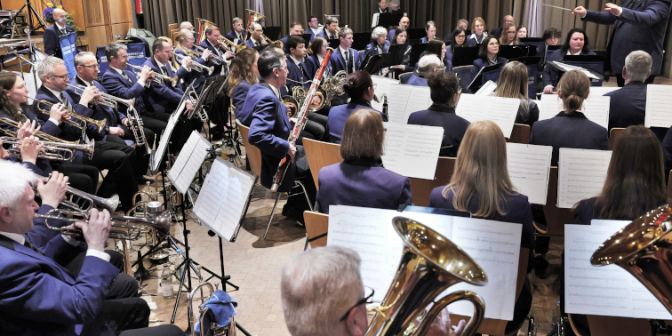 Blasorchester sitzt auf der Bühne und spielt, Dirigent steht vorne und leitet das Konzert.