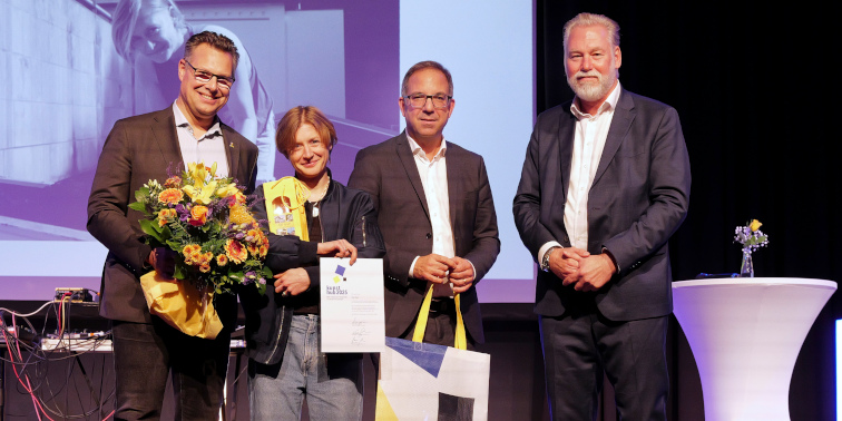 Vier Personen auf einer Bühne bei einer Preisverleihung, eine hält einen Blumenstrauß, eine andere eine Urkunde, alle lächeln in die Kamera.