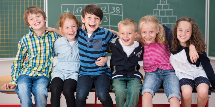 Sechs Schulkinder sitzen auf einem Tisch vor einer Schultafel.