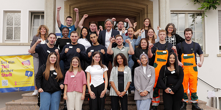 Gruppenbild neuer Azubis vor Rathaus: junge Erwachsene in Alltags- und Arbeitskleidung, lächeln, heben motiviert die Fäuste.