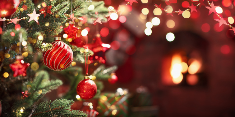 Festlich geschmückter Weihnachtsbaum mit roten Kugeln und Lichtern, im Hintergrund leuchtet ein Kaminfeuer