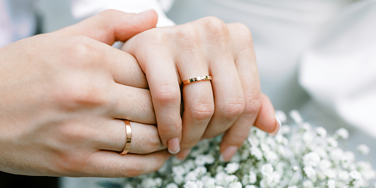 Samstagstrauungen Zwei Hände mit Eheringen, die sich sanft halten, mit weißen Blumen im Hintergrund.