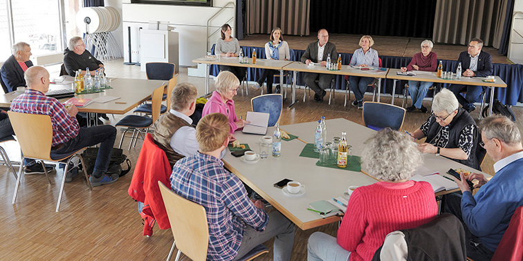 Eine Gruppe von Erwachsenen sitzt in einem Raum um Tische in U-Form. Vorn sitzt ein Podium, die anderen hören zu und machen Notizen.