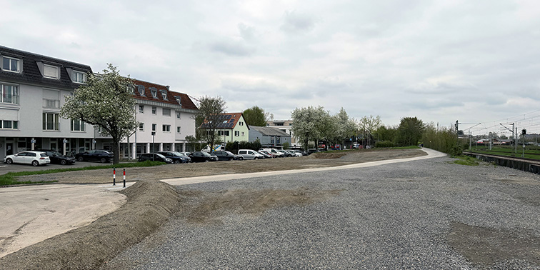 Eine geschotterte Fläche neben Wohnhäusern mit parkenden Autos. Rechts verläuft ein Weg entlang von Bahngleisen, im Hintergrund Bäume und ein bewölkter Himmel.