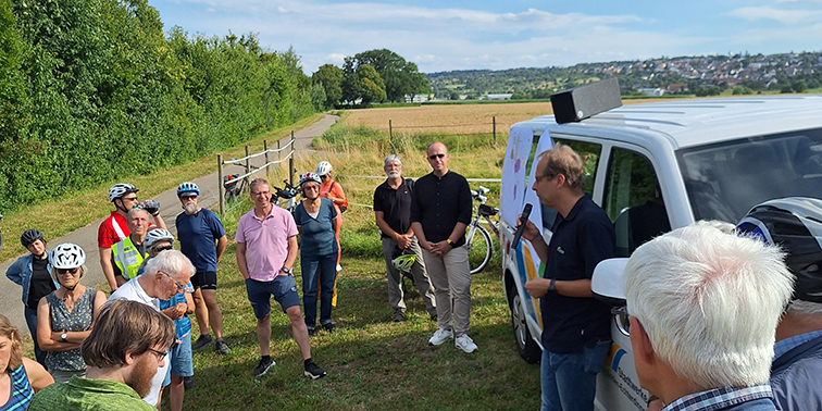 Eine Gruppe von Menschen steht bei sonnigem Wetter auf einer Wiese neben einem weißen Transporter. Ein Mann spricht mit Mikrofon zur Gruppe. Viele Teilnehmer tragen Fahrradhelme, im Hintergrund stehen Fahrräder. Es ist eine organisierte Radtour mit Gesprächsrunde im Freien.