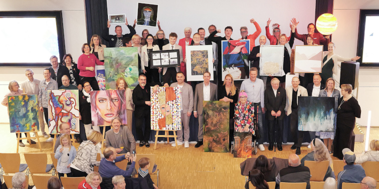 Große Gruppe steht mit Kunstwerken auf einer Bühne, Besucher fotografieren bei einer Vernissage.