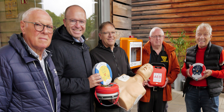 Fünf Männer stehen vor einem Gebäude und zeigen Defibrillator-Geräte sowie eine Übungspuppe – Vorstellung eines neuen Defibrillators.