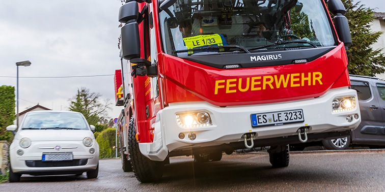 Ein rotes Feuerwehrfahrzeug fährt eine Wohnstraße entlang, im Hintergrund stehen Häuser und parkende Autos.