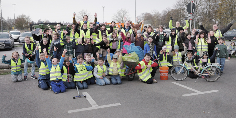 Kinder und Erwachsene in Warnwesten posieren mit Müllsäcken und Werkzeugen bei einer Putzaktion im Freien.