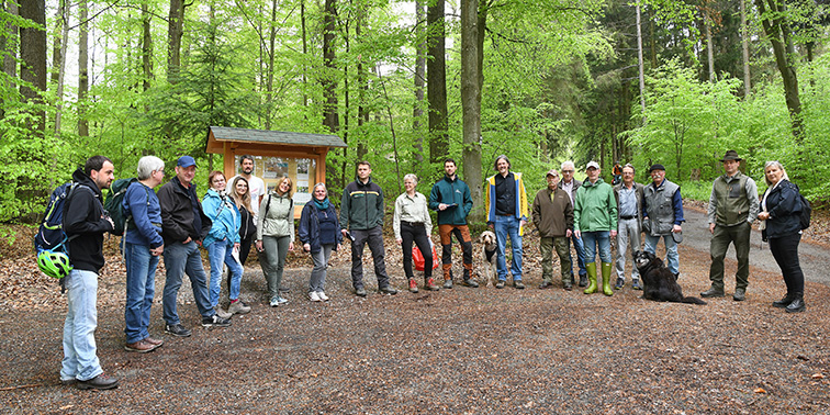 Eine Gruppe von Menschen steht in einem Wald auf einem Waldweg, einige tragen Outdoor-Kleidung, und im Hintergrund ist eine hölzerne Infotafel zu sehen.